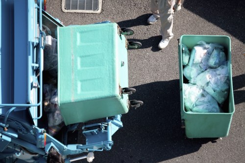 An overhead view of a teal wheeled rubbish bin partially open, revealing mixed waste inside, situated on a paved surface next to a larger teal container filled with discarded plastic bags and packaging materials. Nearby, a person dressed in light clothing is seen standing, with only the lower legs and feet visible, in an outdoor setting possibly related to waste collection or clearance. The scene appears to be part of an urban or suburban environment, highlighting the process of rubbish removal typical for local services in Hackbridge or surrounding areas, with visible textures of the plastic bins and the dark asphalt pavement supporting a professional waste management context. The image emphasizes the collection of general waste as part of a rubbish clearance service provided by Office Clearance Hackbridge, situated in the London Borough of Sutton or nearby, illustrating routine disposal activities.