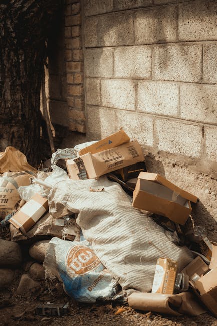 A pile of various waste and cardboard boxes, some partially crushed, resting directly on the ground beside a weathered brick wall and a rough concrete block wall. The cardboard items are of different