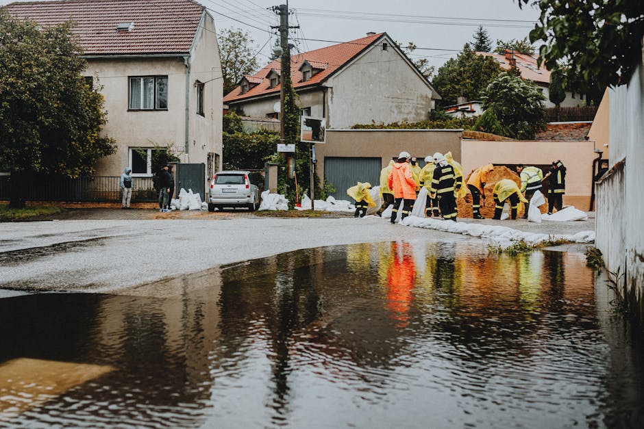 A group of workers dressed in waterproof yellow and orange protective suits and helmets are engaged in clearing flood-damaged debris from a residential street. They are lifting and stacking large whit