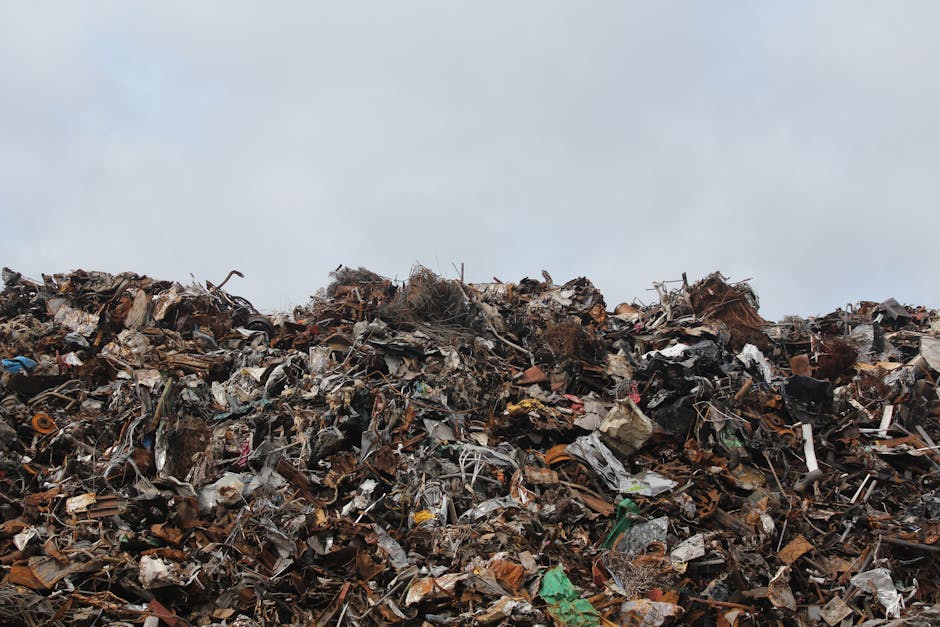 A large pile of assorted waste materials including crushed metal, broken plastic, shredded cardboard, and various discarded household and industrial items, stacked outdoors against a cloudy sky. The w