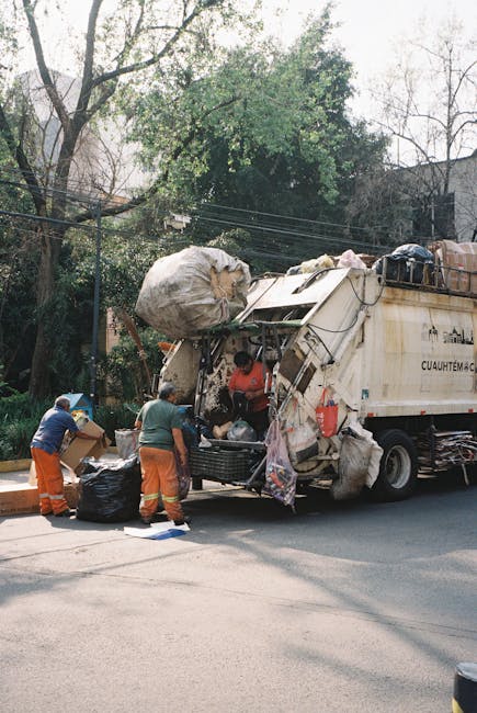 Three workers wearing orange high-visibility trousers and blue shirts are standing beside a large, white rubbish collection truck parked on a street during daytime. The truck’s rear loading area is