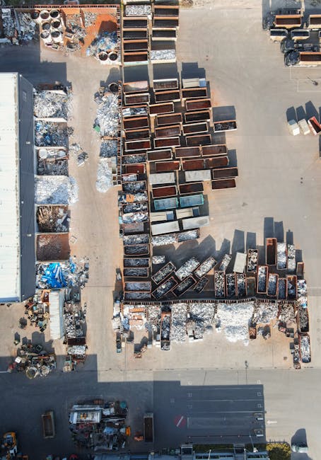 A large industrial scrap metal recycling site featuring a high-reaching mechanical claw suspended from a crane, which is actively picking up a mixture of metallic debris, including twisted and battere