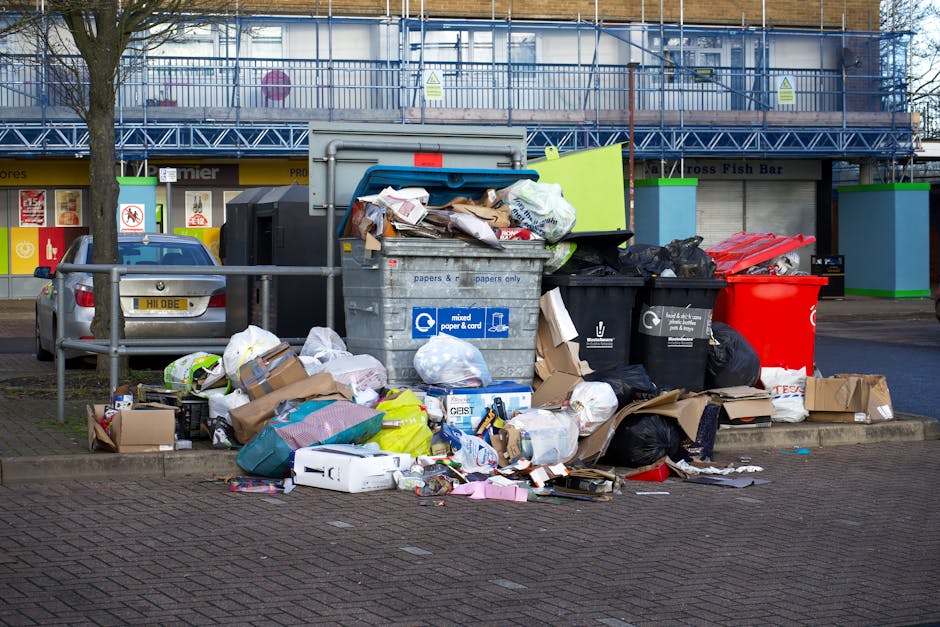 A row of four large, white plastic wheelie bins with closed lids, positioned on a paved surface beside a wooden post, alongside a dark metal shipping container situated in an outdoor area. The bins ar