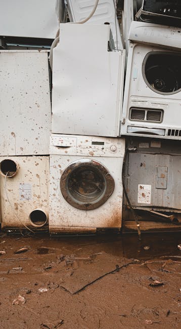A white front-loading washing machine with its door open, revealing the stainless steel drum inside, positioned outdoors against a weathered pinkish wall with visible textures and some rust marks near